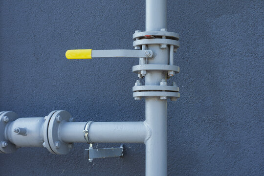 Yellow lever handle gate valve mounted on a gray steel pipe with bolted flanges and union fitting against a textured gray wall background.