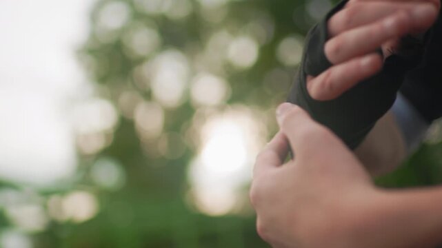 outdoor hands putting on fingerless glove, closeup wrist applying compression wrap for training preparation, soft bokeh background, warm backlight, tactile fingers adjusting thumb loop, subtle motion,