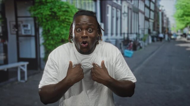 Man pointing to self with both hands raised on city street in daylight amid row of buildings; surprise doubt guilt.