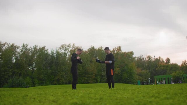 Outdoor green field two white men coach and trainee practice sparring prep, exchanging guidance and gloves near tree line under cloudy sky, calm reflective mood, mentorship and measured warmup, wide