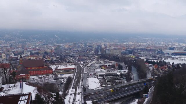 Aerial view of Innsbruck, Austria, shows railyard and station, highway bridge over the Inn River, red historic complex, snow dust, low clouds, and light traffic movement.