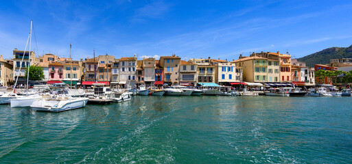 Obraz premium Panaorama du port de Cassis, avec yachts et voiliers amarrés devant les maisons colorées du front de mer et les collines verdoyantes sous un ciel bleu, Var, Provence, France
