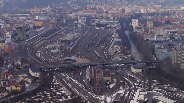 Aerial view of Innsbruck, Austria, showing rail yard, station, curved tracks, freight cars, road bridge, Inn River, church spires, and snow in muted winter light.