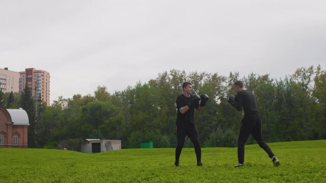 two men boxing drills near buildings with close pad work and jabs, coach and student dynamic, apartment blocks and church dome in background, green lawn, cloudy sky, technical focus and persistence