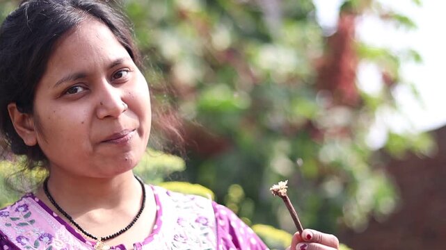 Young Indian woman using a traditional babool datun stick for oral hygiene outdoors, showcasing natural dental care and rural lifestyle.