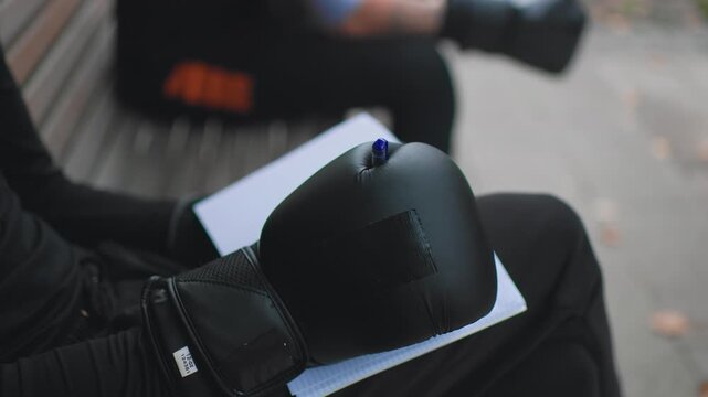 boxing glove with pen on notebook closeup, fighter lap, ink mark on page, journal used for training notes and tactic sketches, textured leather surface, outdoor bench, quiet preparation