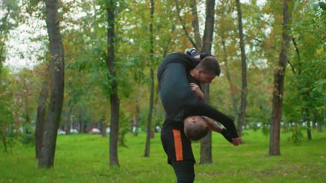park takedown and ground struggle, aggressive clash escalates as one man lifts and slams opponent onto path, hands reaching, shoes scuffing, visible strain, no bystanders, dense tree backdrop