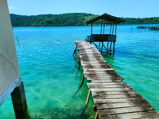 A weathered wooden pier extends to a small stilt hut over stunning turquoise water, backed by a...
