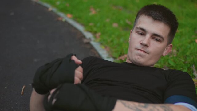 caucasian man fighter lying on grass beside pathway, catching breath and checking gloves with focused gaze black training top, visible tattoo, lone cooldown moment in park with autumn tones,