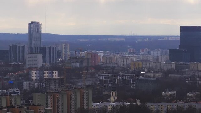 Aerial daytime view over Katowice, Poland shows mid century blocks, newer towers, a tall mast, cranes by an emerging skyscraper, and distant chimneys in haze.