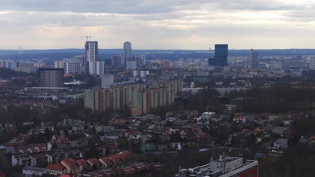 Aerial sweep over Katowice, Poland shows modern high rises, a tower under construction with crane, antennas, red roof houses, leafless parkland, overcast late winter light.