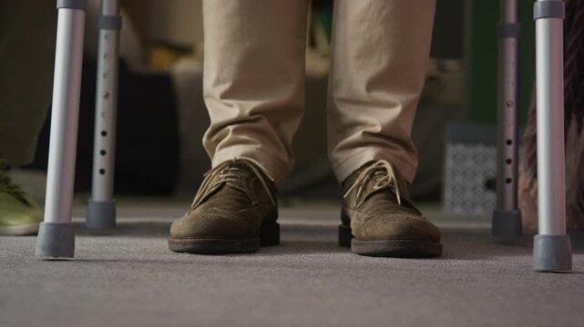 Close-up low section shot of unknown man wearing beige pants and brown shoes slowly moving towards camera with help of walking frame