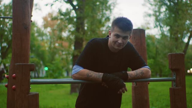 man resting on parallel bar smiling, late afternoon park scene with wooden posts and metal rail, relaxed posture with visible tattoos and gloves, friendly expression, postworkout recovery vibe