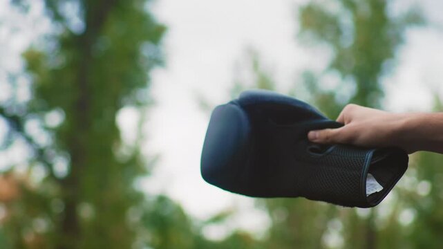 Compact speaker held outdoors near park trees, Side view of meshcovered portable speaker by leafy trees, Vlogger demonstrating small outdoor speaker against backdrop of park greenery