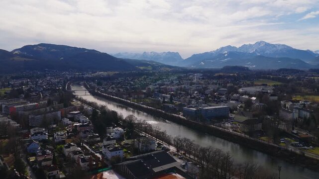Aerial drone shot follows the Salzach River in Salzburg, Austria, past bridges, hospitals, and apartments, as a smooth forward descent reveals Alpine peaks in winter haze.