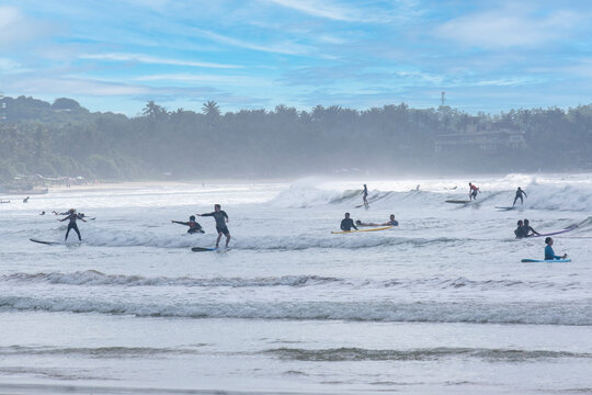 8.12.2025. Weligama Sri Lanka. Crowded surf spot in Weligama bay, Sri Lanka. Group of young people and beginners learning to surf with boards on the beach and in the Indian ocean waves.