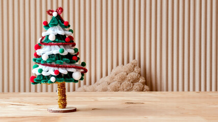 A decorative Christmas tree ornament stands on a wooden table, against a backdrop with a textured pattern. It is adorned with colorful pom-poms, embellishments and a star on top. © Stock4u2024 