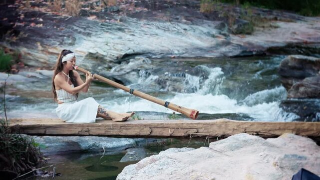 A Serene Performance: A Woman Playing a Bamboo Instrument didgeridoo While Seated on a Log Over a Tranquil Stream Surrounded by Nature's Beauty
