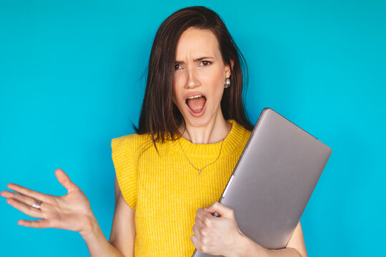 Angry and furious young woman screaming while holding closed laptop, gesturing with irritation and frustration, feeling outraged by work problems or tech failure, isolated on blue background