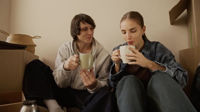 Medium shot of young Caucasian man and woman sitting on floor among packed boxes drinking tea and talking in bright apartment during moving day