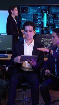 Diverse team of software developers and engineers discussing data analytics in a high-tech office. A man holds a tablet while collaborating near screens with UI code and mobile interfaces