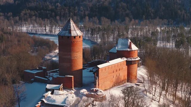 Aerial drone circles Turaida Castle in Sigulda, Latvia, showing red brick towers, cylindrical keep, crumbling walls, snow roofs, icy Gauja River, long low light shadows.