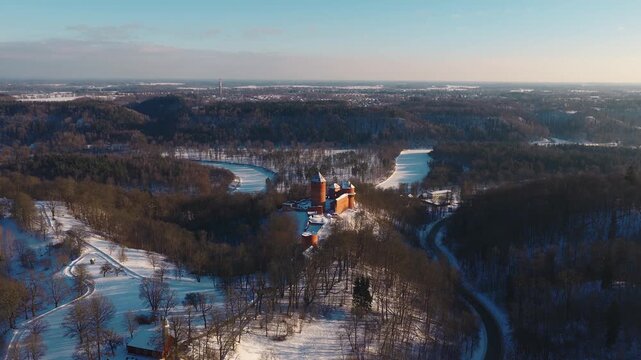 Aerial view shows Turaida Castle on a wooded hill in Sigulda, Latvia, the frozen Gauja River, snow parkland, bare trees, and a winding road as the drone glides slowly.