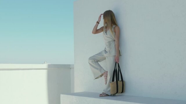 Stylish woman posing in light linen outfit with straw bag against white wall