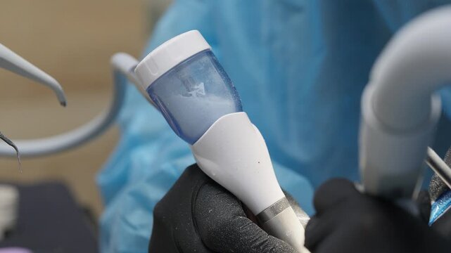 Close-up of dental air polishing handpiece with powder container in dentist's gloved hands during professional teeth cleaning procedure in modern dental clinic