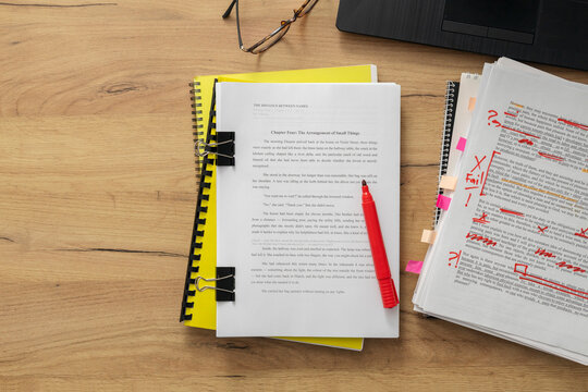 Top view of a cluttered editor's desk during a document review session. Printed pages with red ink markups, binder clips, markers, and notebooks reflect the detailed process of professional copy