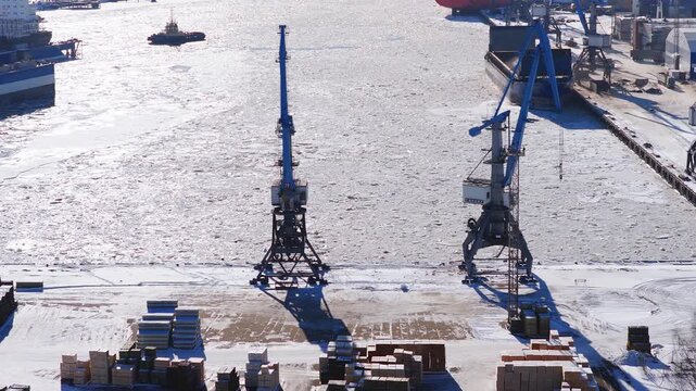 Aerial view of Riga Shipyard in winter shows crane, red ship, floating drydock, stacked containers, and a tugboat moving through icy water under sharp sunlight.