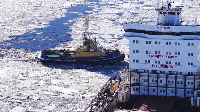 Aerial view shows Riga Shipyard in Latvia as tug SFINKSA maneuvers beside a white ship with red SAFETY FIRST and NO SMOKING, IMO 9448671, through ice under crisp daylight.