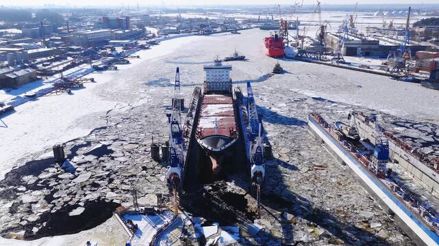 Aerial Riga Shipyard shows a dry dock with a bulk carrier hull, blue gantry cranes, repair quays, and a red tanker in an icy channel as tugboats maneuver at midday.
