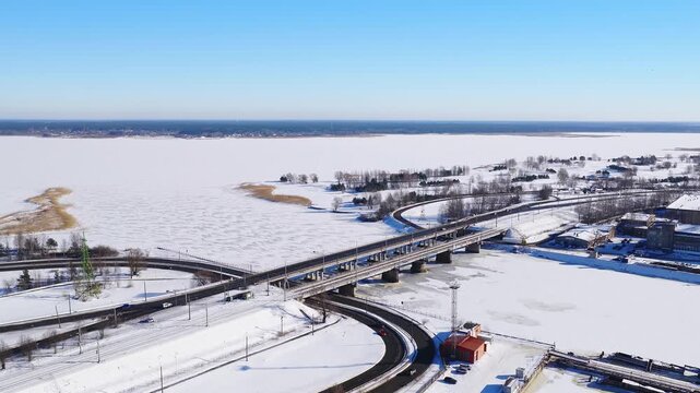 Aerial view shows Riga Shipyard in Riga, Latvia, a multi span concrete bridge, looping ramps, rail lines, reed islets, icy Mezaparka lake, cars and trains in bright winter sun.