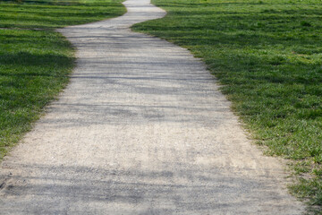 Obraz premium A light colored winding gravel walkway curves through a lush green field of grass in a sunny outdoor park