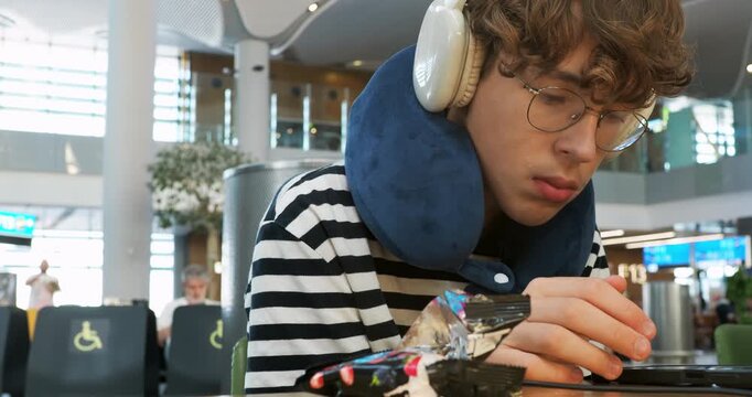 Young man with headphones and neck pillow sits tiredly in an airport waiting area. He looks at his phone while waiting for a flight. Modern travel terminal background with other passengers