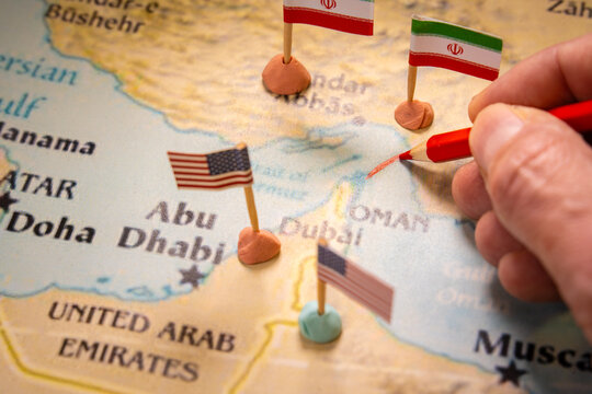 Conceptual image of a hand drawing a red line on a map of the Middle East with United States and Iran flags marking the Strait of Hormuz. Represents geopolitical conflict and global oil supply crisis.