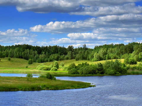 A panoramic view of a peaceful lake in the Polish Lake District surrounded by dense green forest. The calm water surface and white clouds create a perfect summer landscape in North-Eastern Poland.