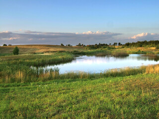 Fototapeta premium A lush green meadow and pond under a evening summer sky in the rural Masurian region. Distant forests create a scenic pastoral view of the Polish countryside.