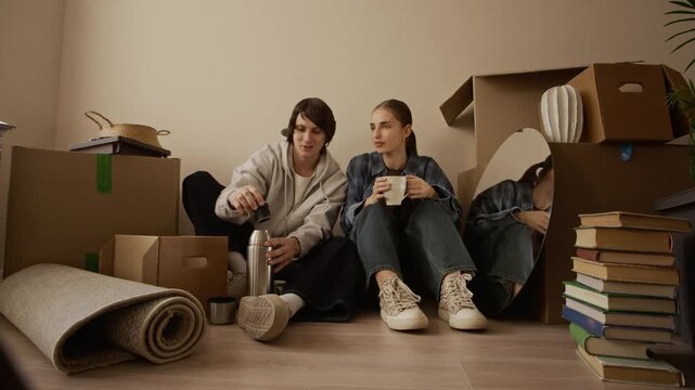 Young couple sitting on floor among cardboard boxes drinking tea from thermos and chatting after moving into bright new apartment, eye level wide shot showing relocation and home settling moment