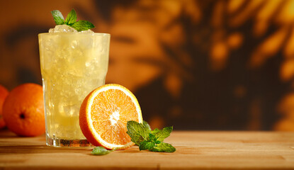 Cold orange fruit drink placed on a rustic wooden table with soft, moody lighting. The background is blurred, featuring a warm interior and a wall with subtle leaf shadows, creating a cozy and elegant © magdal3na