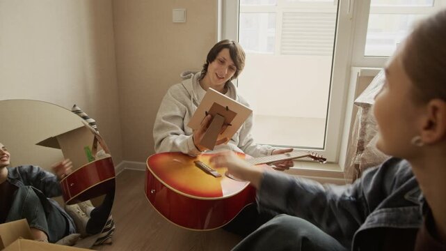 Medium eye level shot of young Caucasian couple smiling while looking at framed photo of their dog during unpacking belongings from cardboard boxes in bright cozy living room of new apartment