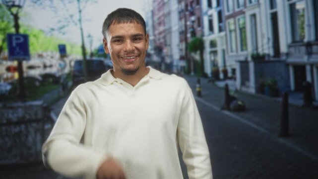 Man pointing both index fingers down while smiling, wearing a cream sweater in front of row houses on a street; playful confidence.