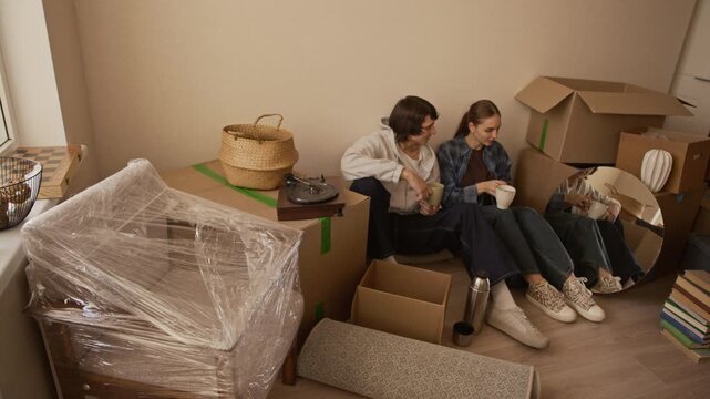 High angle wide shot of young Caucasian couple taking break while sitting among cardboard boxes and household belongings in new apartment, drinking tea from thermos during moving day