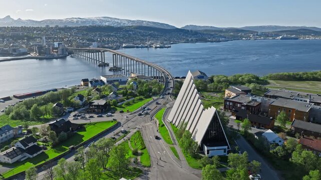 Aerial view circling the Arctic Cathedral in Tromsdalen, Tromso, Norway. Modern landmark church sits beside the road and fjord with Troms&oslash; Bridge stretching across the water.