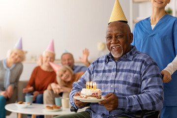 Fototapeta premium A man in a wheelchair smiles while holding a birthday cake with candles. Friends and caregivers surround him, wearing party hats. The event takes place indoors in a bright room.