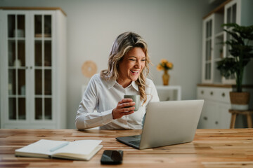 Happy woman enjoying remote work from home, holding a coffee mug and laughing while looking at her...