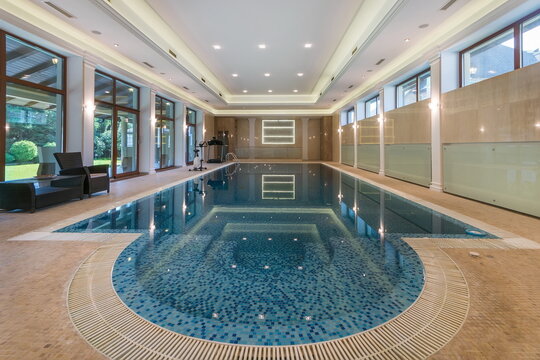 An indoor swimming pool is seen in a private residence. The pool has clear blue water and is surrounded by large windows. Sunlight fills the space