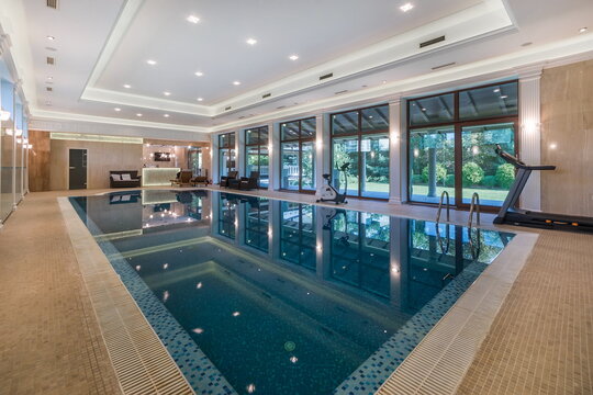 A spacious indoor swimming pool is seen next to a fitness area. Natural light fills the room, highlighting the clear water. Exercise equipment is placed near the pool