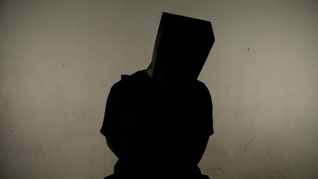 An anonymous man with a bag over his head sits strapped to a chair in a dark room.